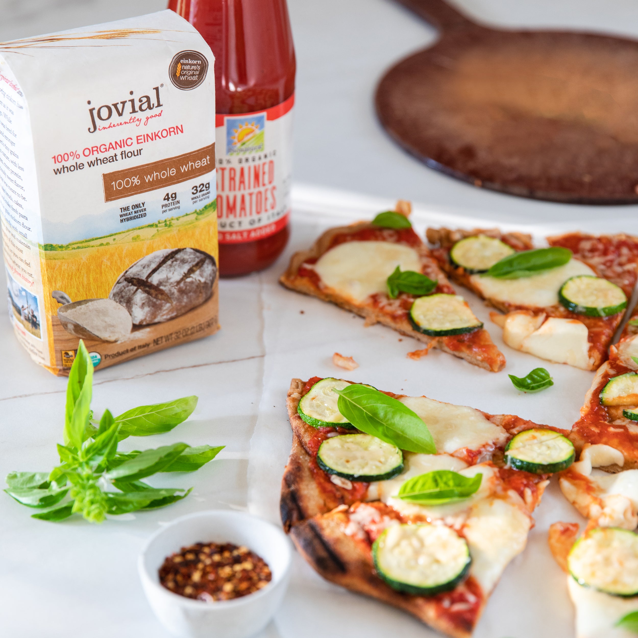 Jovial organic einkorn whole wheat flour bag beside sliced whole-wheat pizza with zucchini, mozzarella, and basil