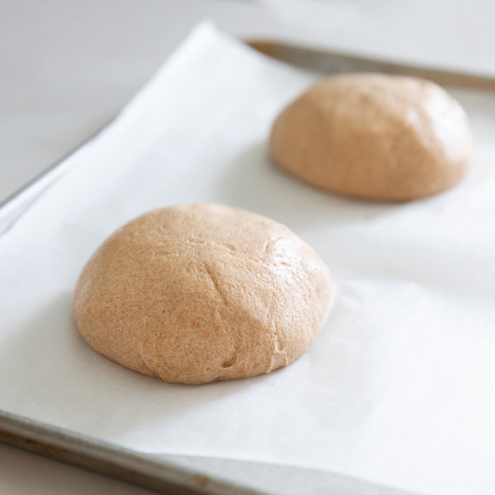 Close up of two smooth, risen balls of whole wheat einkorn pizza dough resting on a parchment-lined baking sheet
