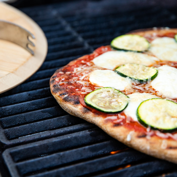 Close-up of whole-wheat einkorn pizza on a grill, charred crust with tomato sauce, melted mozzarella, and zucchini slices