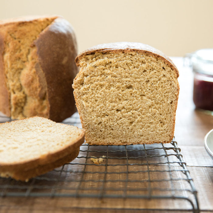 Front view of a sliced Einkorn whole wheat sandwich loaf showcasing soft, airy crumb and golden crust on a cooling rack with jar of jam in the background