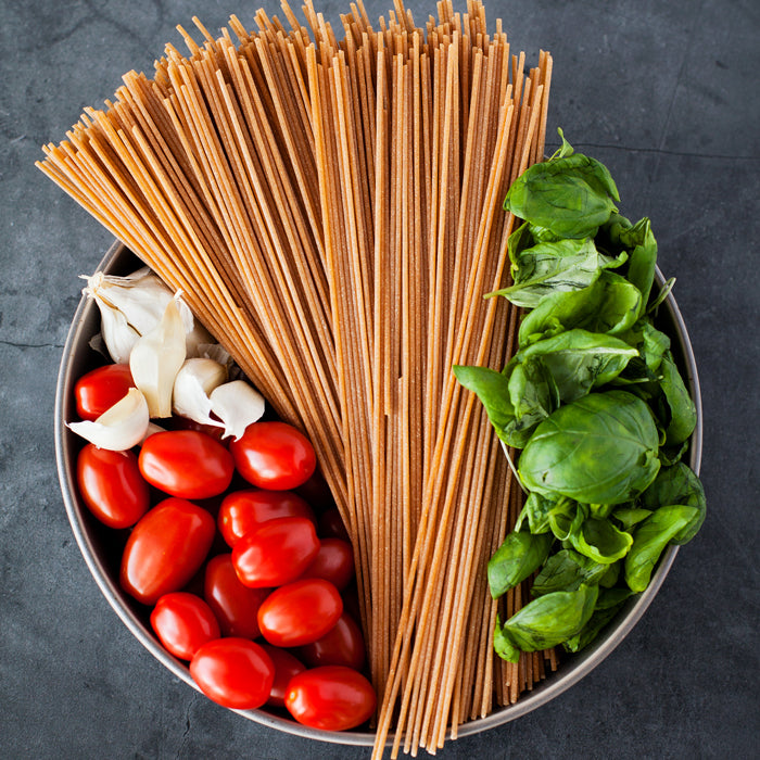 Top-down view of whole wheat spaghetti with cherry tomatoes, garlic, and basil in a metal bowl on dark slate