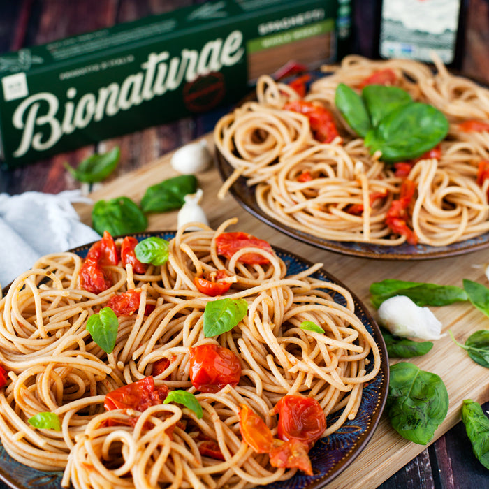 Two plates of whole wheat spaghetti with roasted cherry tomatoes and fresh basil on a wooden board, Bionaturae pasta box in background