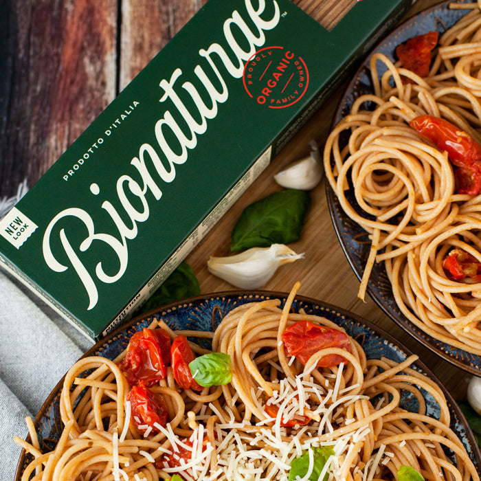 Two bowls of whole wheat spaghetti alla Checca with roasted cherry tomatoes, basil, grated cheese and a Bionaturae pasta box on a wooden surface