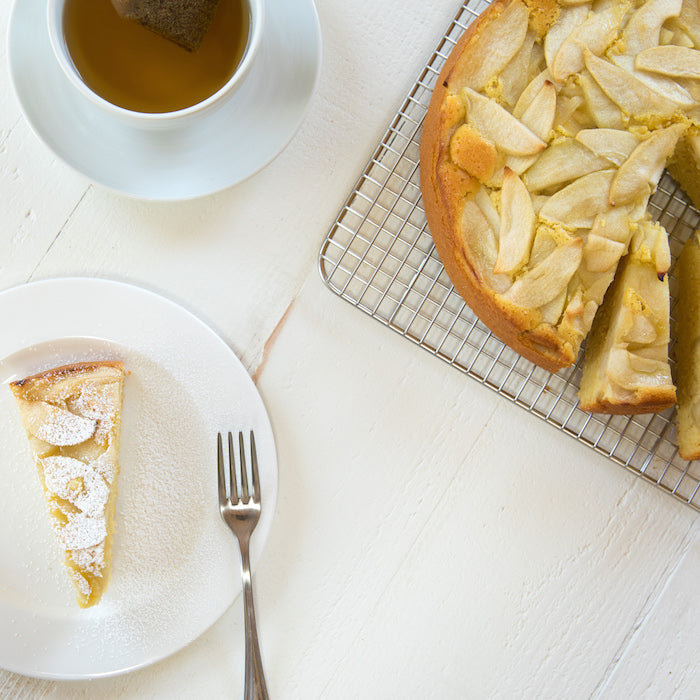Overhead of an einkorn apple cake: a powdered-sugar-dusted slice on a plate with fork, whole apple-topped cake and tea