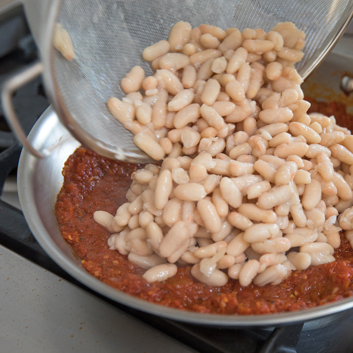 Hands pouring drained cannellini beans from a colander into tomato sauce in a skillet on the stove