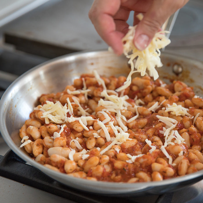 Person sprinkling shredded cheese over tomato-simmered cannellini beans in a stainless-steel skillet on a stovetop