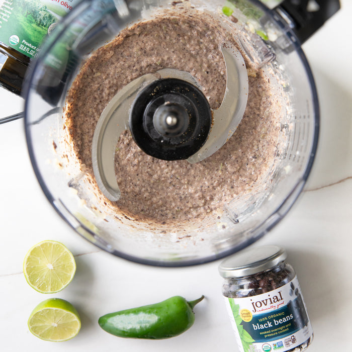 Overhead of blended black bean dip in a food processor with a jar of Jovial organic black beans, halved lime, and a jalapeño