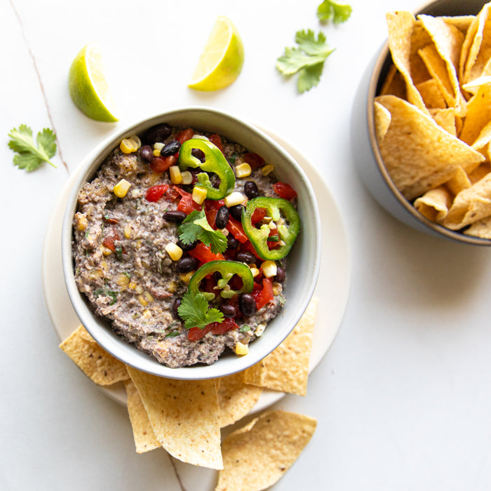 Overhead view of black bean dip topped with corn, tomato, jalapeño and cilantro, with tortilla chips and lime wedges