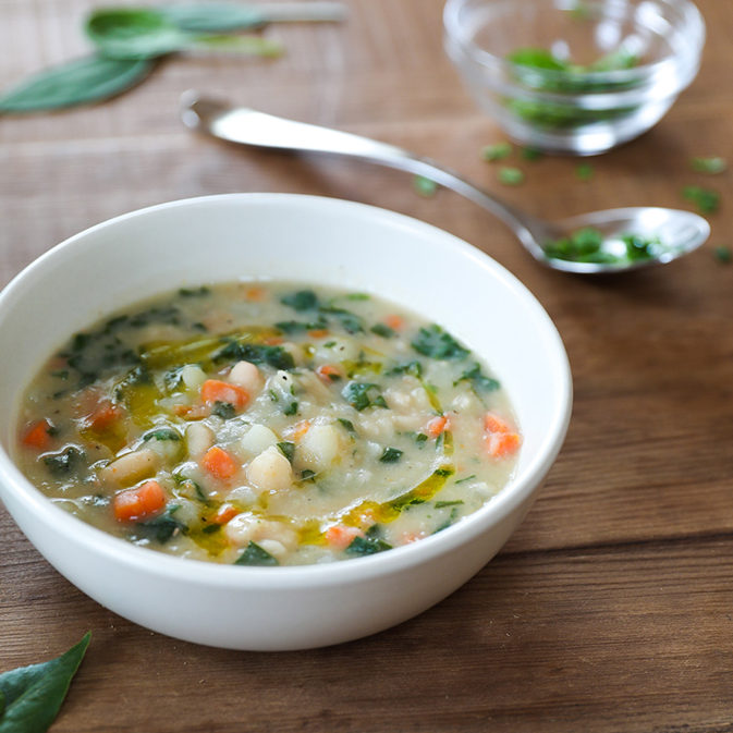 Organic cannellini and potato vegetable soup in white bowl with olive oil drizzle and chopped spinach on wooden table