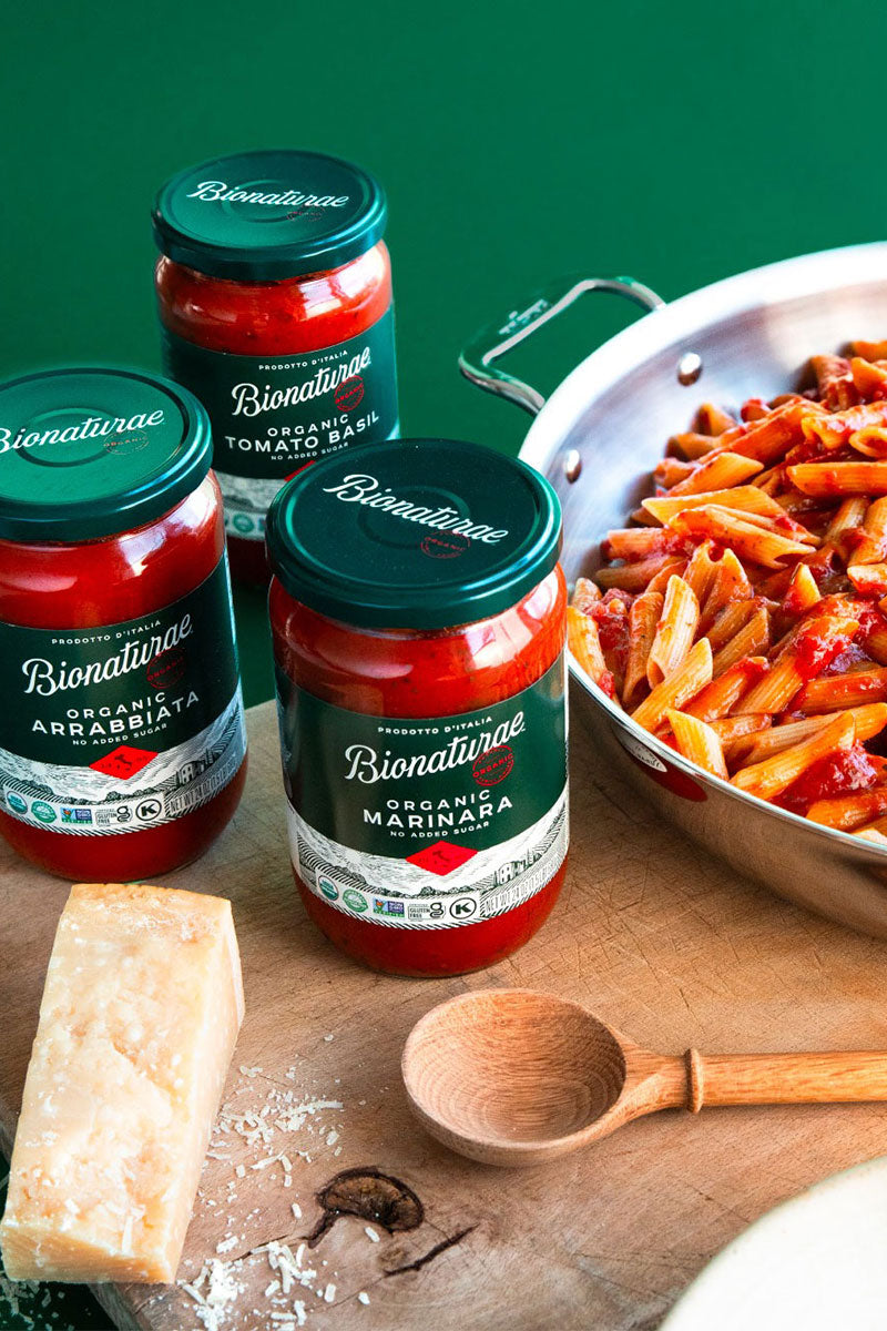 Three Bionaturae organic sauce jars (Marinara, Arrabbiata, Tomato Basil) on board with penne, cheese wedge, wooden spoon