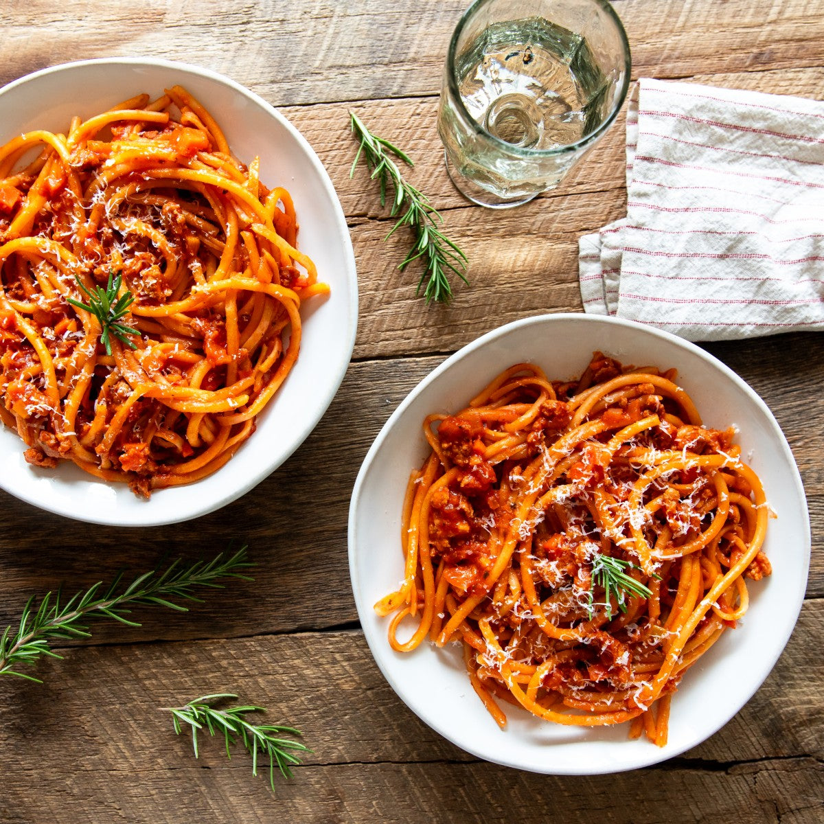 Two bowls of organic gluten-free brown rice bucatini with sausage ragu, grated cheese and rosemary on a wooden table