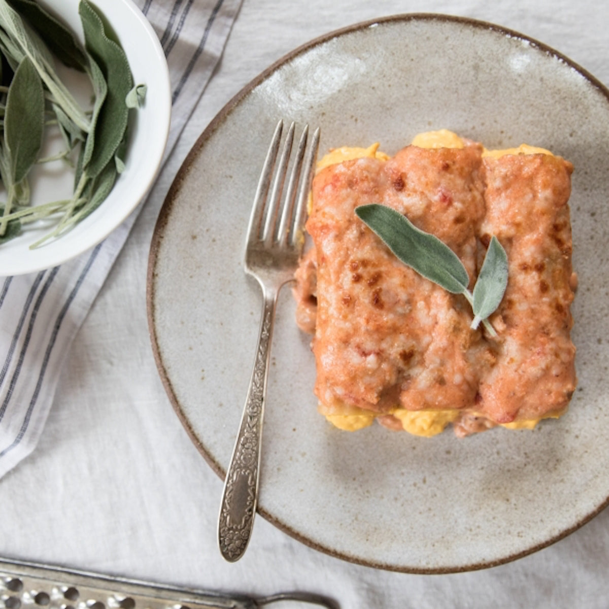 Plated baked organic brown rice manicotti with orange tomato-based sauce and a sage leaf, fork beside on a ceramic plate