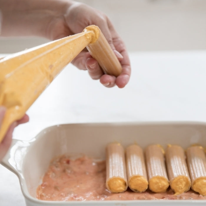 Hands filling gluten-free manicotti pasta with butternut squash filling over tomato sauce in a ceramic baking dish
