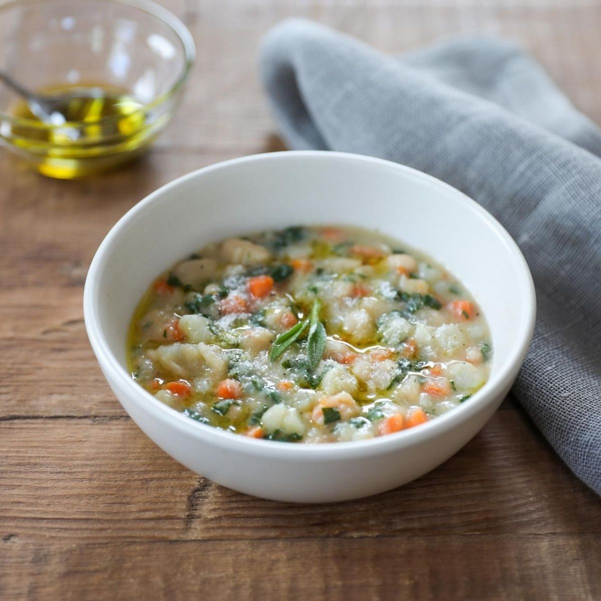 Bowl of organic cannellini bean soup with carrots and greens on a wooden table, bowl of olive oil and gray napkin nearby