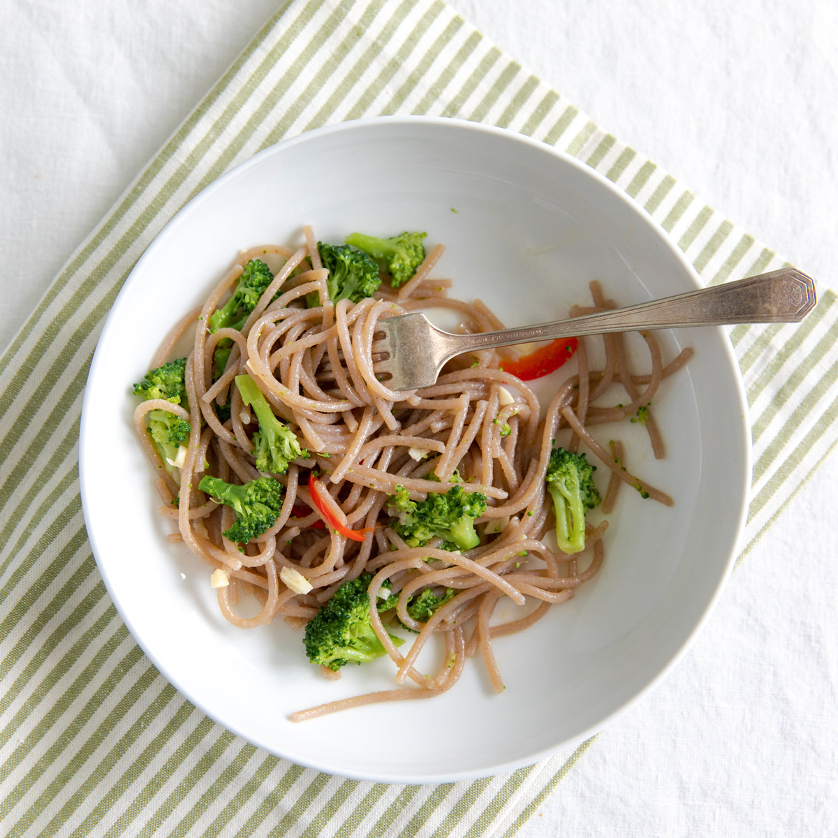 Bowl of cooked organic cassava spaghetti with broccoli and red pepper, fork resting on the bowl over a striped napkin