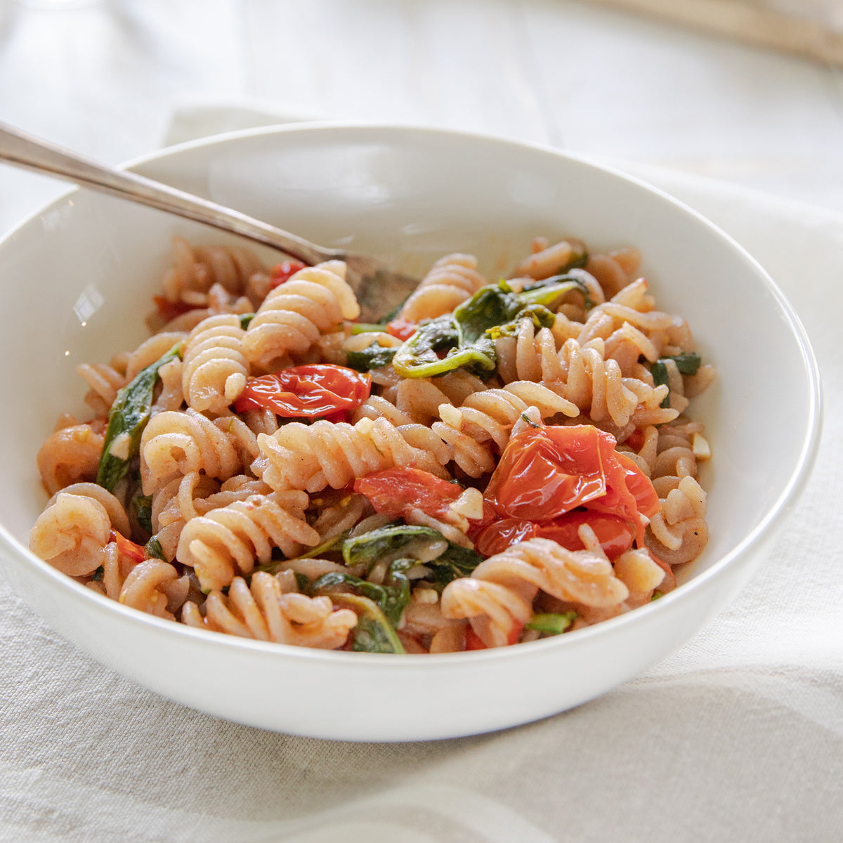 Bowl of cooked organic cassava fusilli with cherry tomatoes and leafy greens on a light cloth backdrop