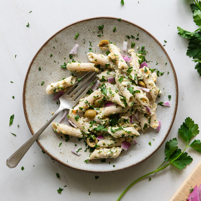 Top-down view of vegan chickpea tuna-style pasta salad showcasing brown rice penne, chopped red onion, green olives, parsley, and a fork on a ceramic plate