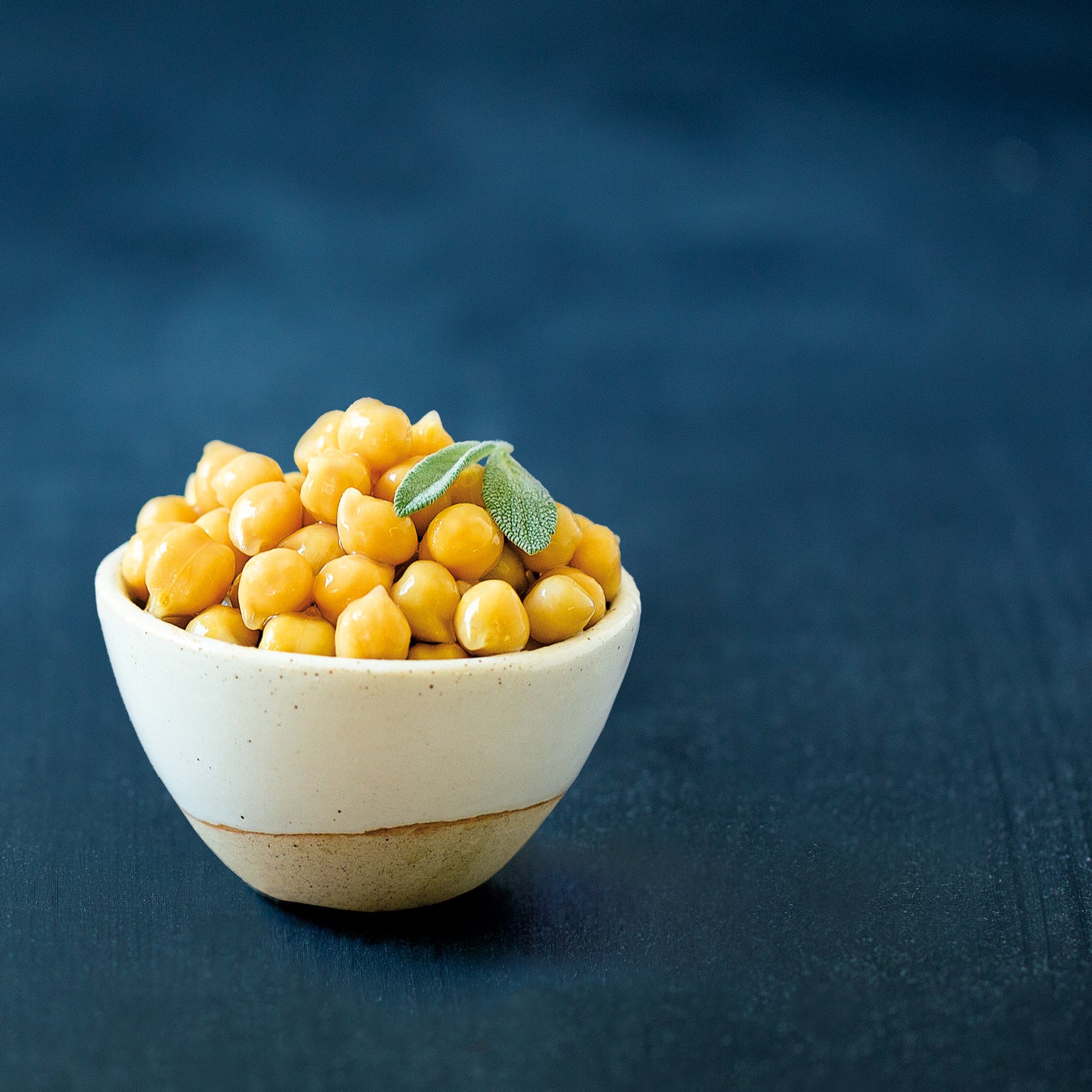 Close-up of cooked organic chickpeas in a small white bowl with a basil leaf on a dark textured background