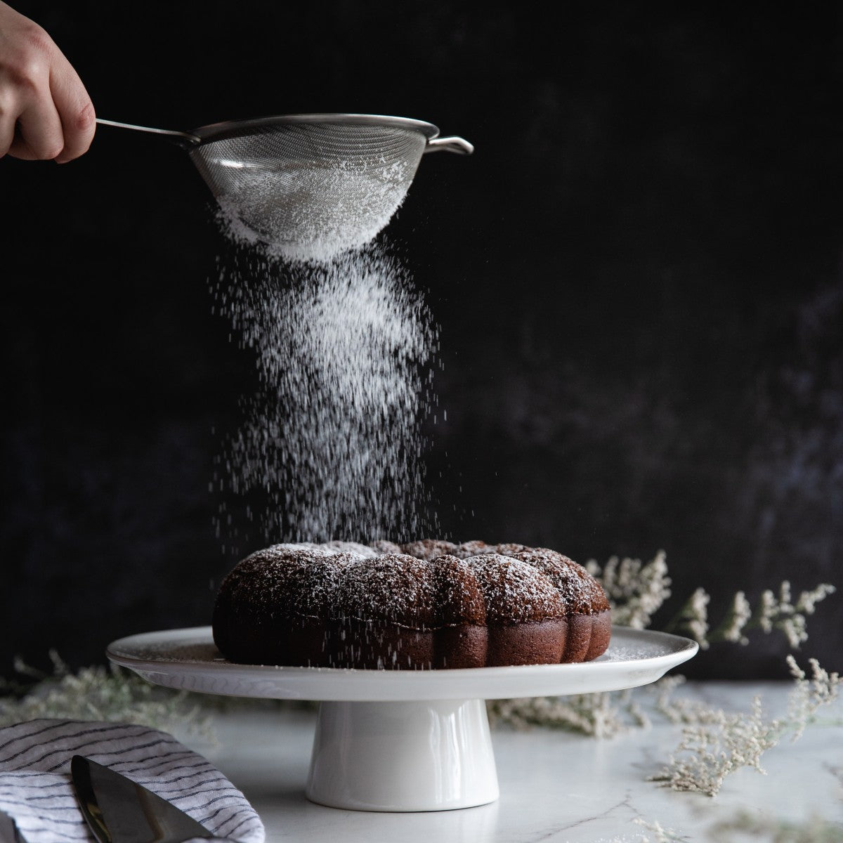 Chocolate olive oil cake being dusted with powdered sugar on a white cake stand against a dark background