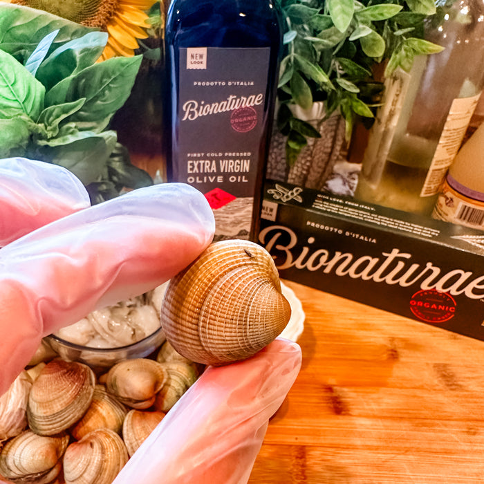 Gloved hand holding a raw clam with Bionaturae extra virgin olive oil bottle, basil, and more clams on a wooden board