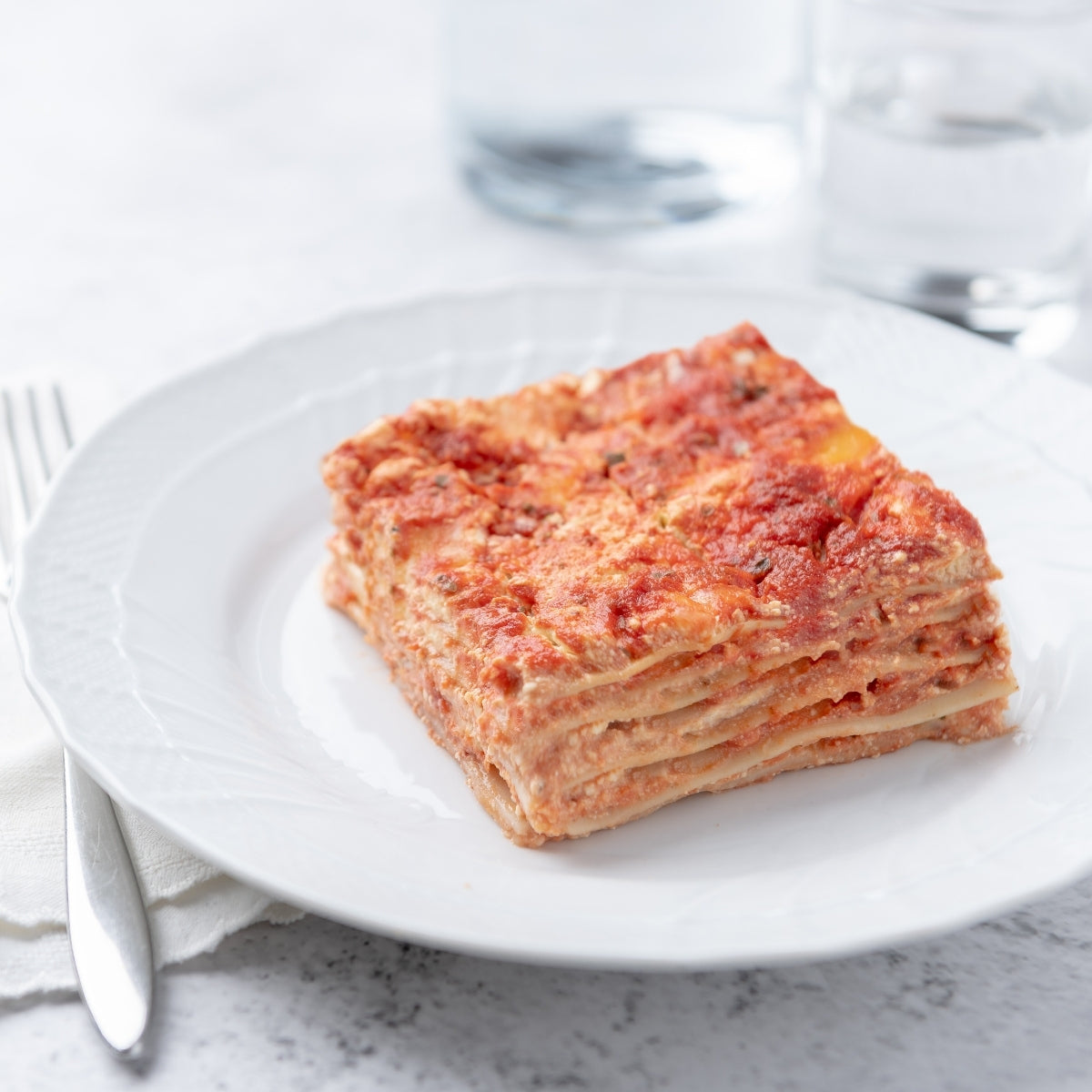 Three-quarter view of Organic Gluten Free Brown Rice Classic Lasagna slice showing layered pasta and tomato sauce on a white plate with fork and water glass