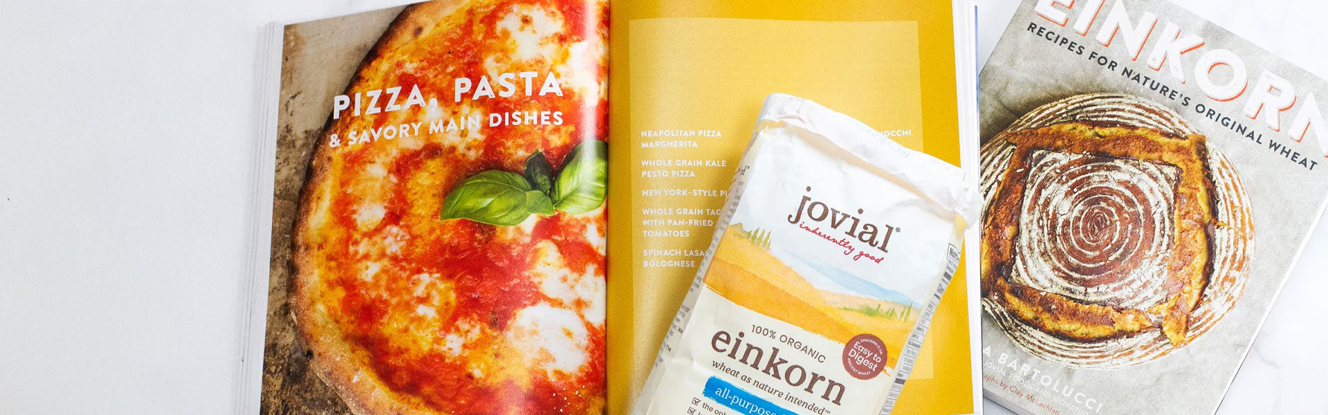 Open Einkorn cookbook showcasing pizza and savory recipes beside a 100 percent organic einkorn flour bag