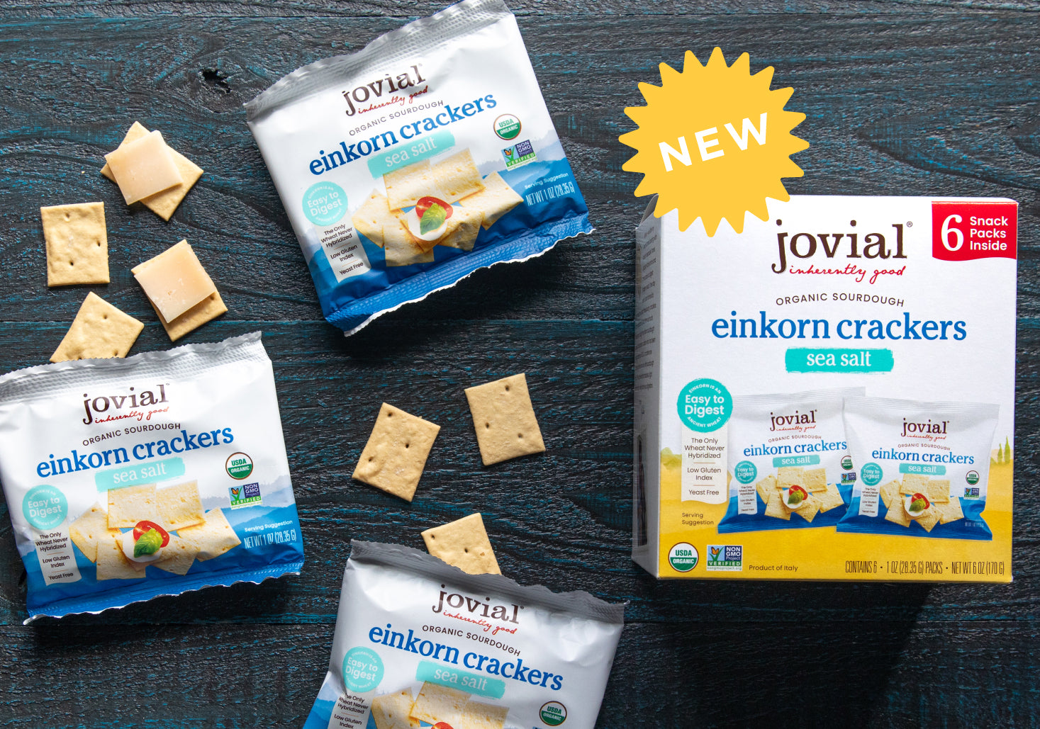 Top-down view of Jovial einkorn crackers sea salt mini snack packs and retail box with yellow NEW burst on dark wood background