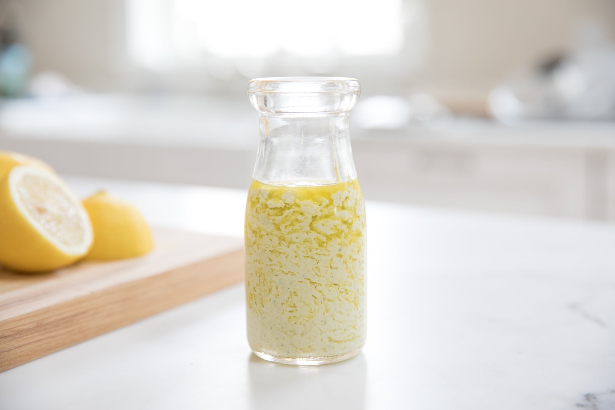Front view of creamy herb olive oil dressing in a glass jar showing emulsified texture with lemon halves on a cutting board