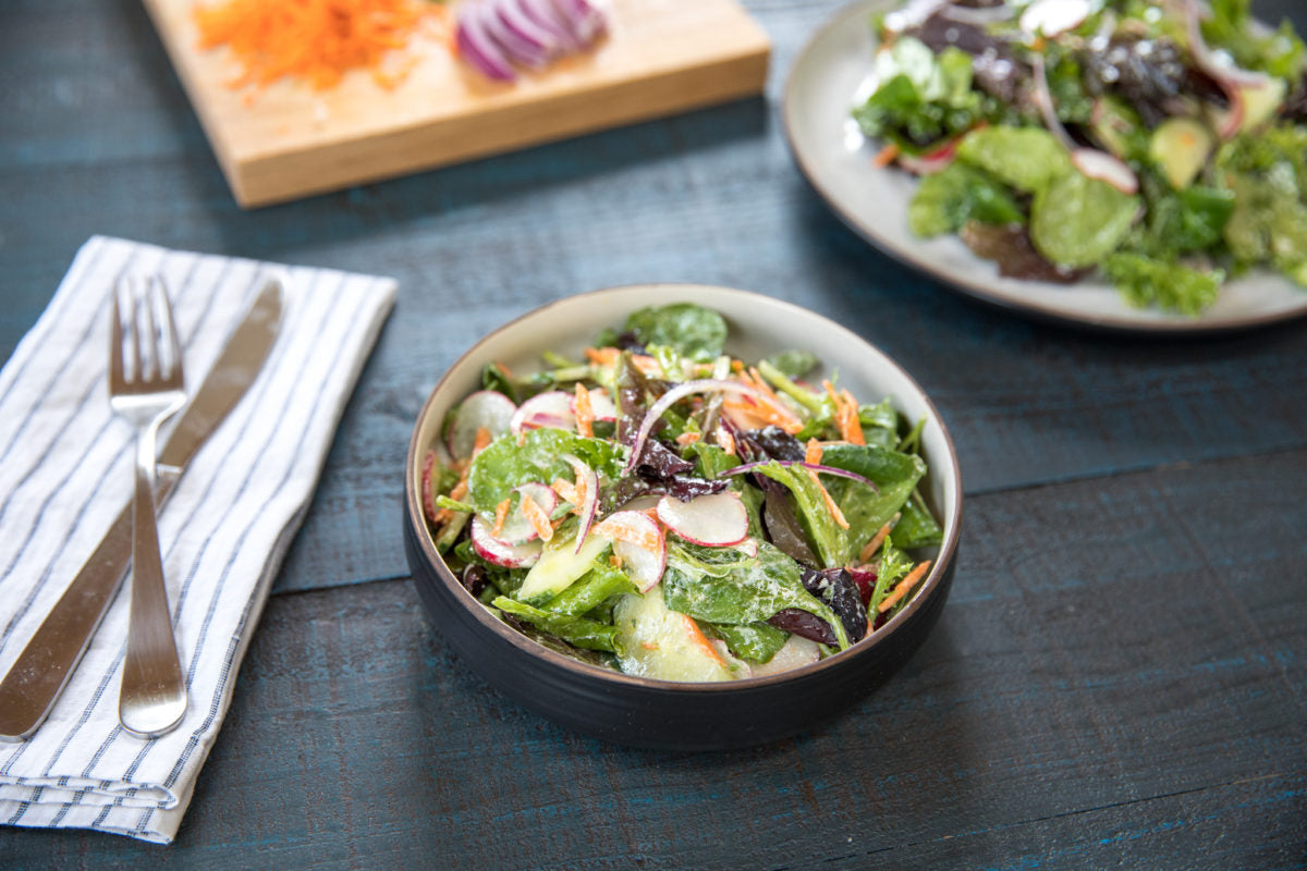 Angled view of mixed greens with creamy herb olive oil dressing, radishes, cucumber and shredded carrot in bowl on dark wood