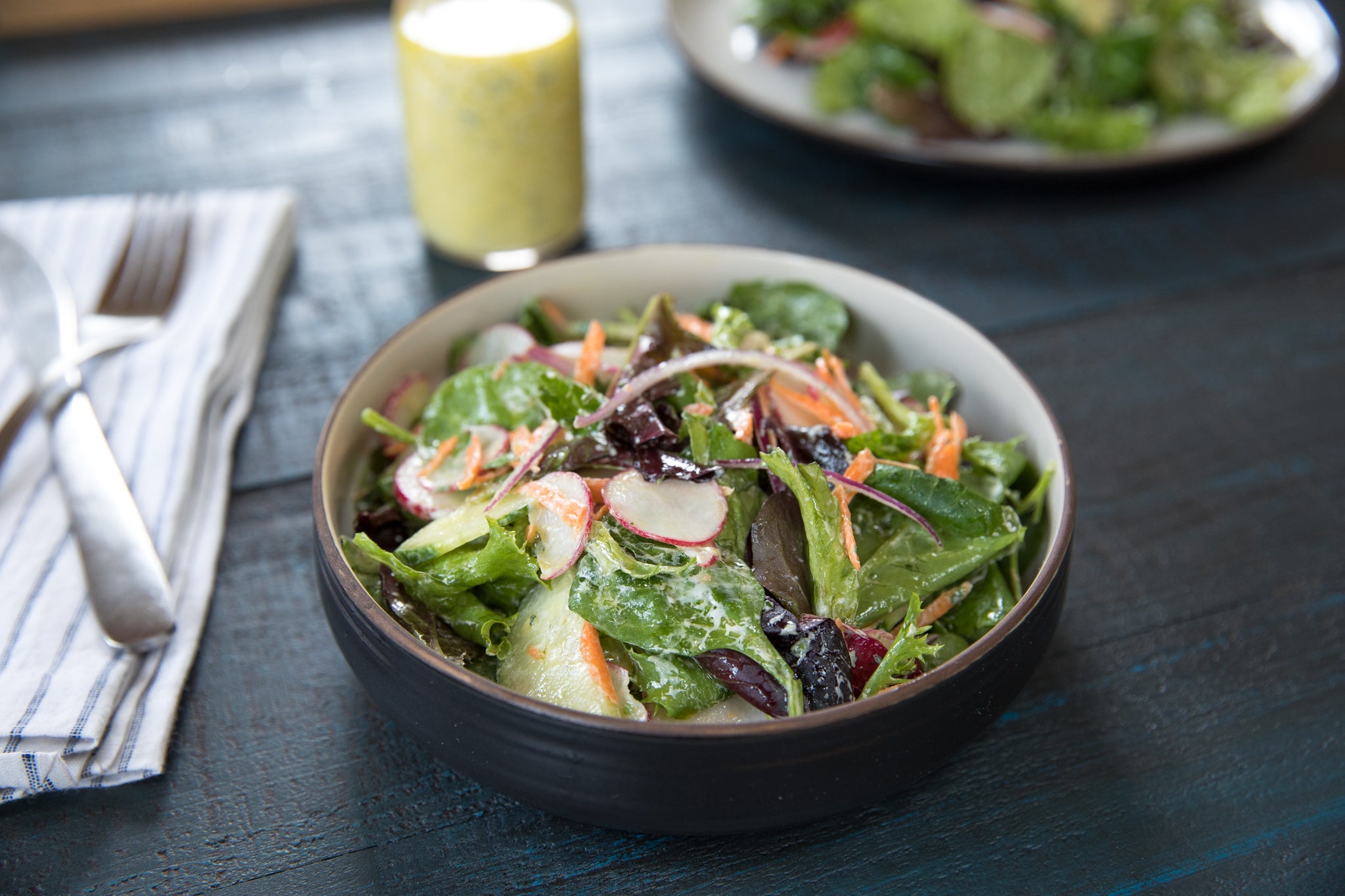 Bowl of mixed green salad with radish and carrot, tossed in creamy herb olive oil dressing on a dark wooden table
