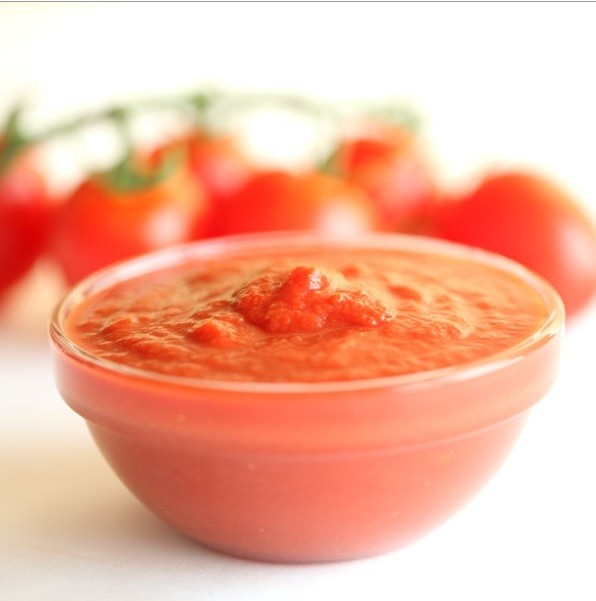 Close-up of organic crushed tomatoes in a glass bowl showing thick texture with ripe tomatoes blurred in the background
