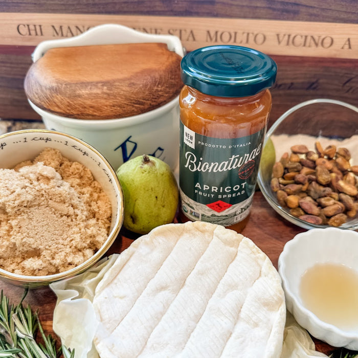 Close-up of Bionaturae Apricot Fruit Spread jar beside a round Brie, pear, pistachios, and brown sugar on a wooden board