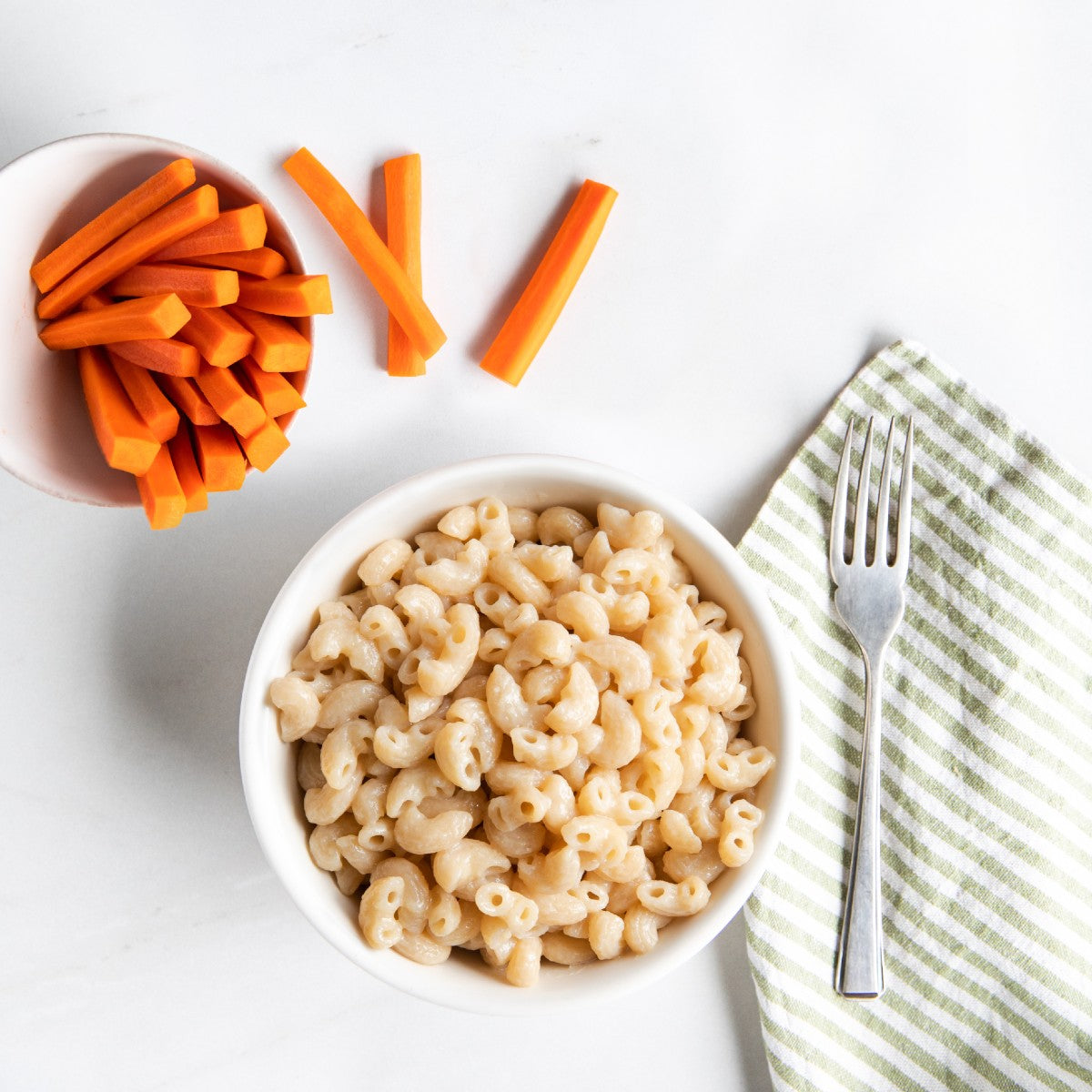 Overhead view of Organic Gluten Free vegan mac in a white bowl showing creamy elbow pasta with carrot sticks and a fork on a striped napkin