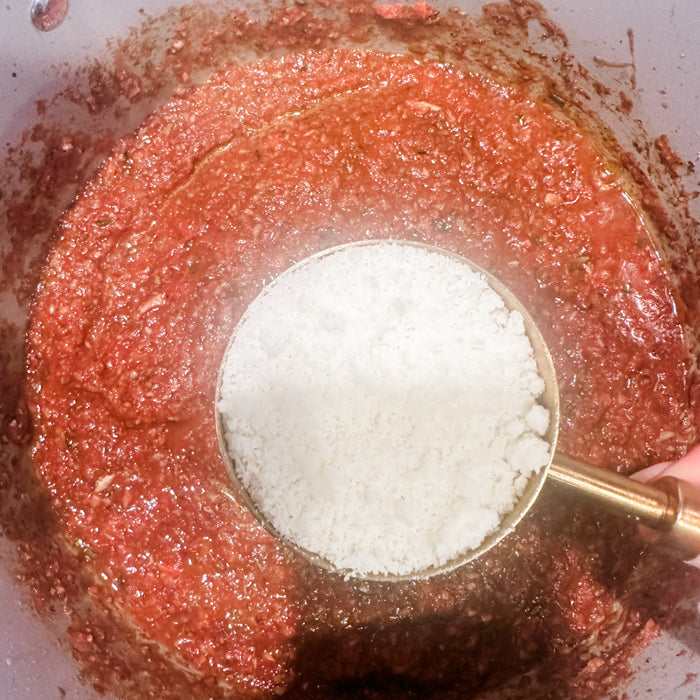 Measuring scoop of grated cheese added to meatless mushroom bolognese sauce in a pot