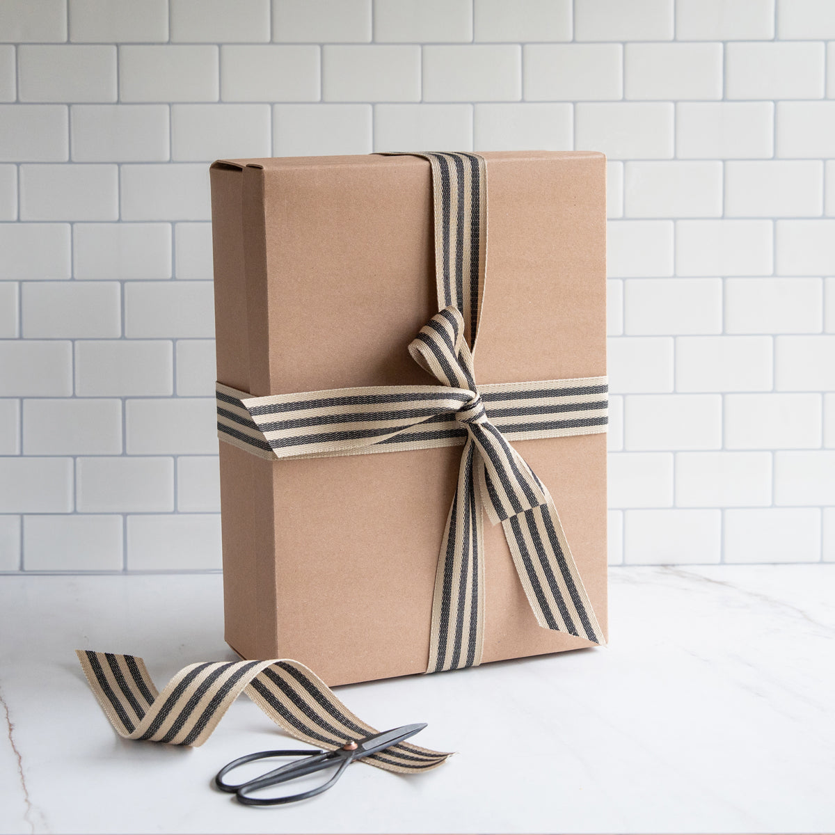 Front view of Premium Einkorn Baking Kit box showcasing kraft gift box tied with black and cream striped ribbon on white tiled background