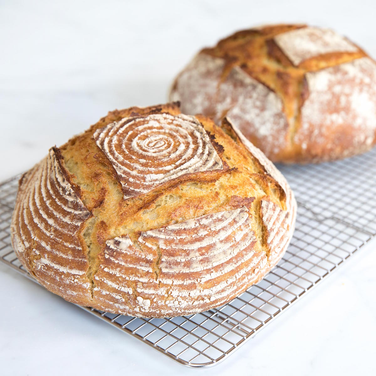 Two round einkorn boules cooling on a wire rack, golden crust with scoring and light flour dusting on a white surface