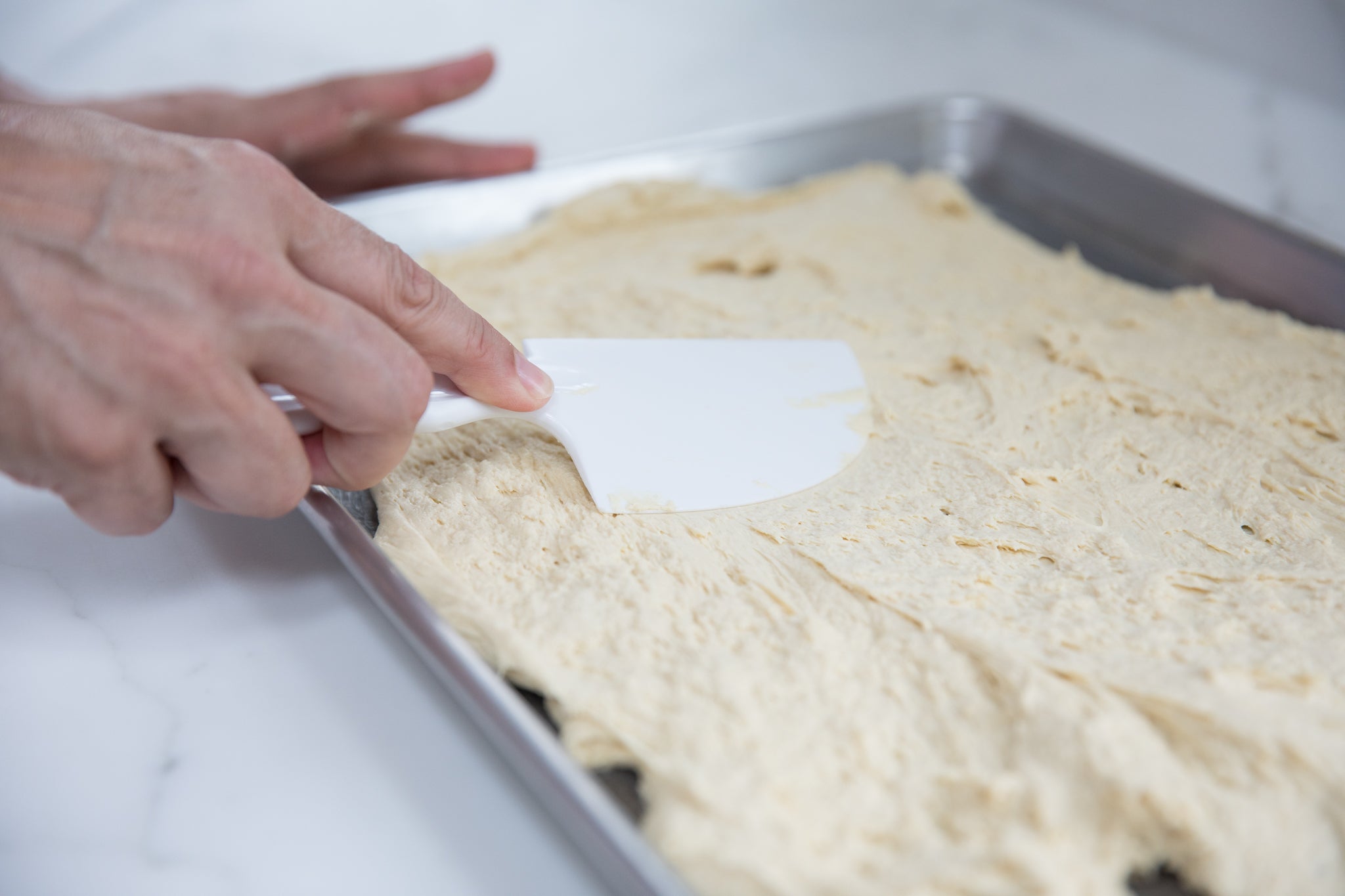 Hands spreading einkorn dough with a white spatula on a rimmed baking sheet
