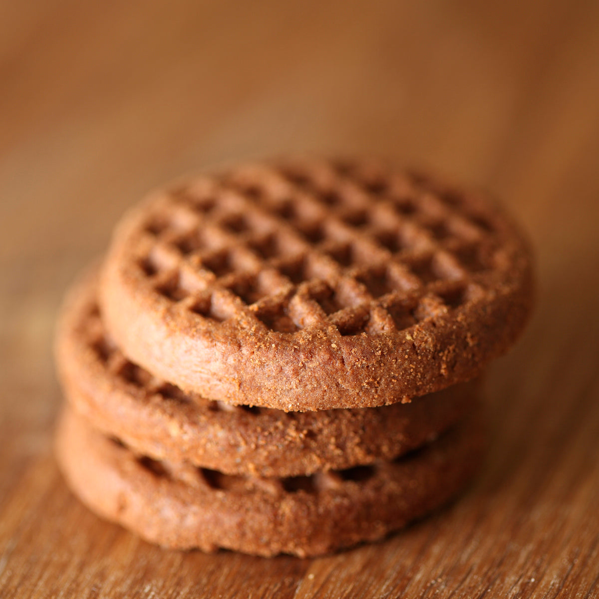 Close-up of Organic Einkorn Crispy Cocoa cookies stacked, showing crisp chocolate texture with blurred wooden background