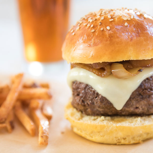 Close-up of Einkorn sesame hamburger bun with beef, melted cheese and caramelized onions, fries and beer behind