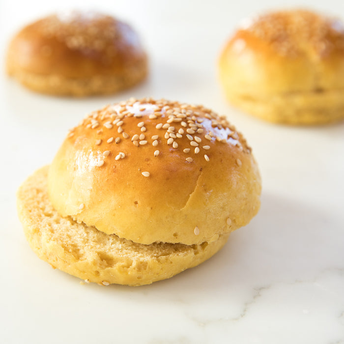 Front view of Einkorn hamburger bun split to show soft interior and sesame-seed topped golden crust on a white marble surface