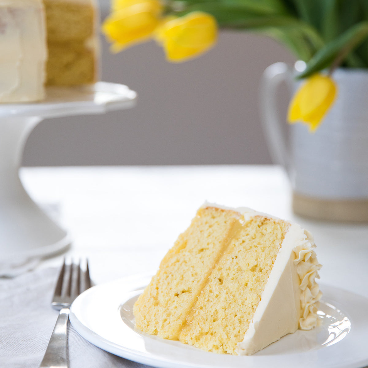 Slice of lemon cake made with organic einkorn flour on a plate with a fork, cake and yellow tulips soft focus behind