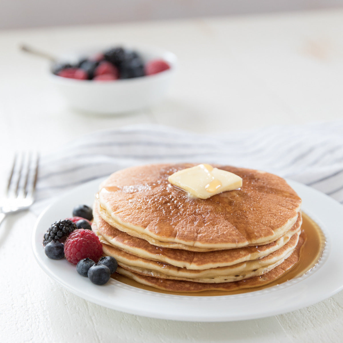 Stack of pancakes topped with a pat of butter, maple syrup, and mixed berries on a white plate
