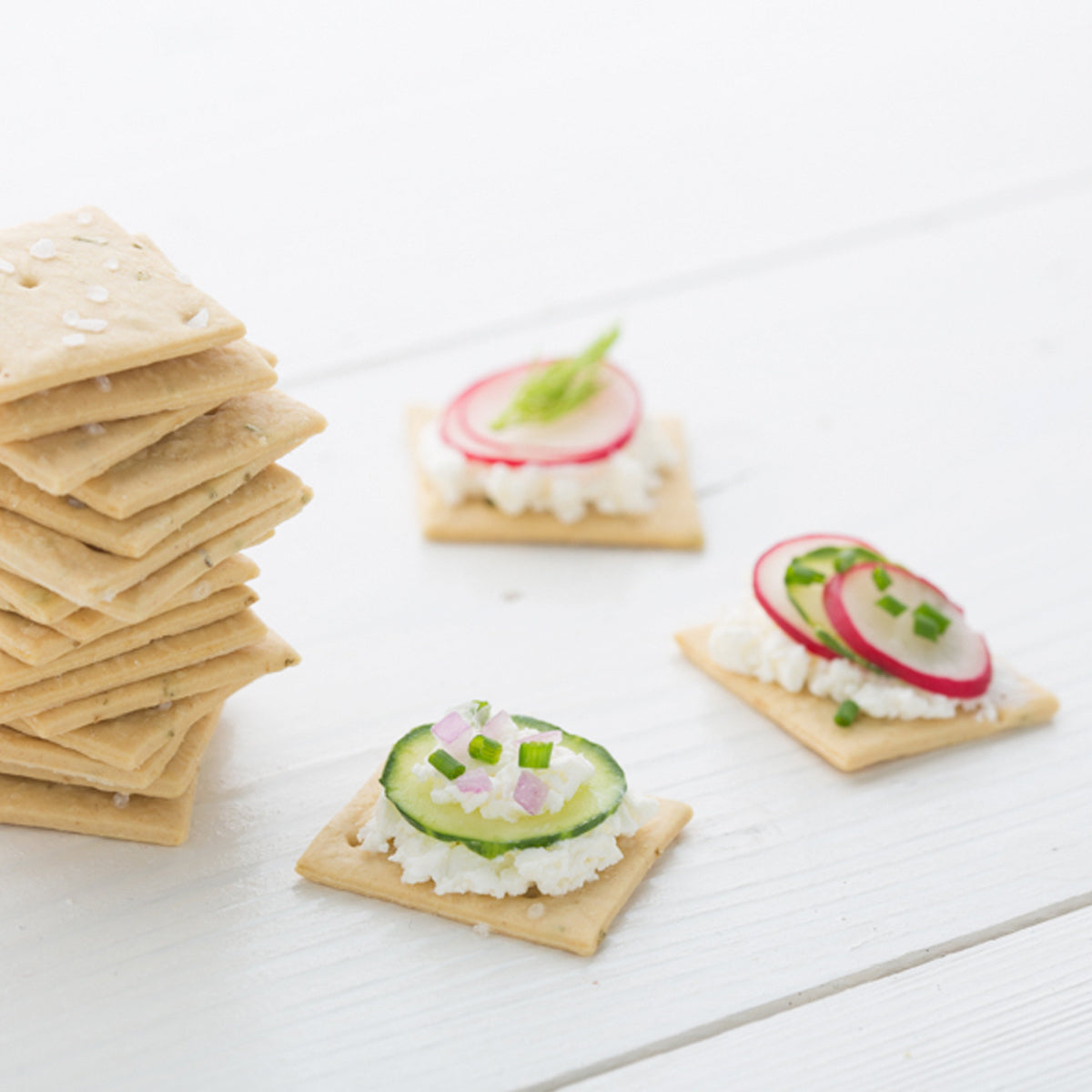 Three-quarter view of Organic Einkorn rosemary sourdough crackers showing a stack and three topped with cucumber and radish