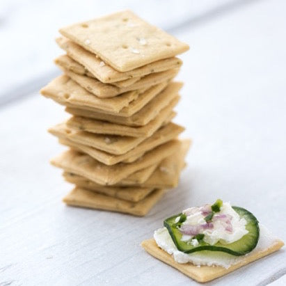 Front view of Organic Einkorn Sourdough Sea Salt Crackers showing a stack and one cracker topped with cucumber on light gray