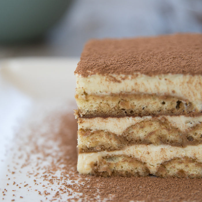 Close-up view of an Einkorn tiramisu slice showing layered ladyfingers and mascarpone dusted with cocoa on a white plate