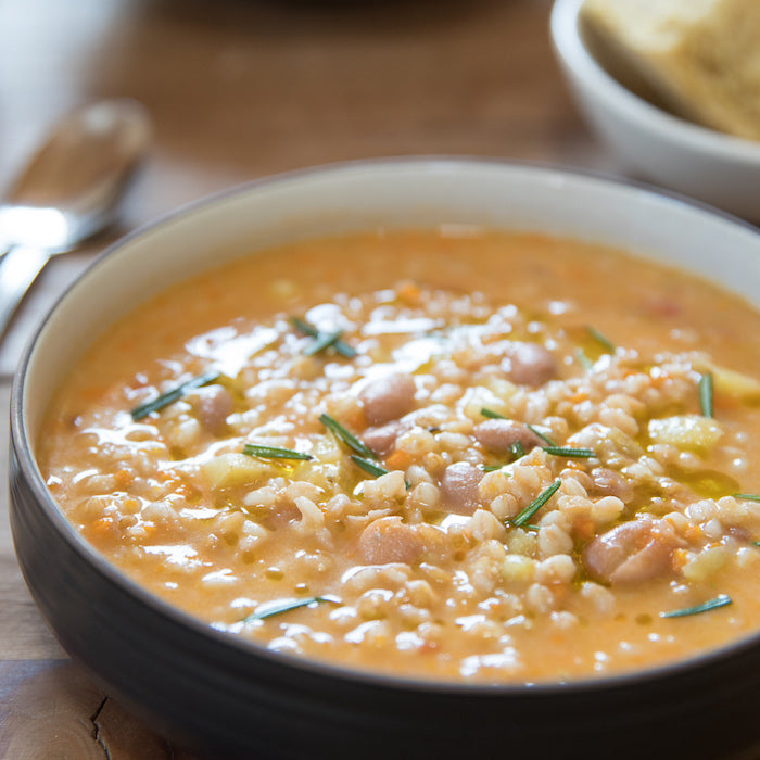 Close up bowl of hearty farro and white bean soup garnished with rosemary and a drizzle of olive oil on a wooden table