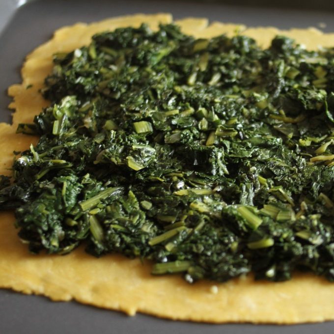 Sautéed mixed greens spread over a rolled einkorn pie crust on a dark baking sheet