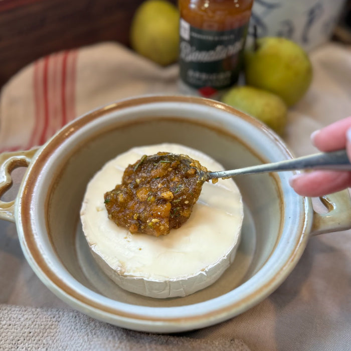 Wheel of Brie being topped with organic apricot fruit spread in a small baking dish, jar and pears in background