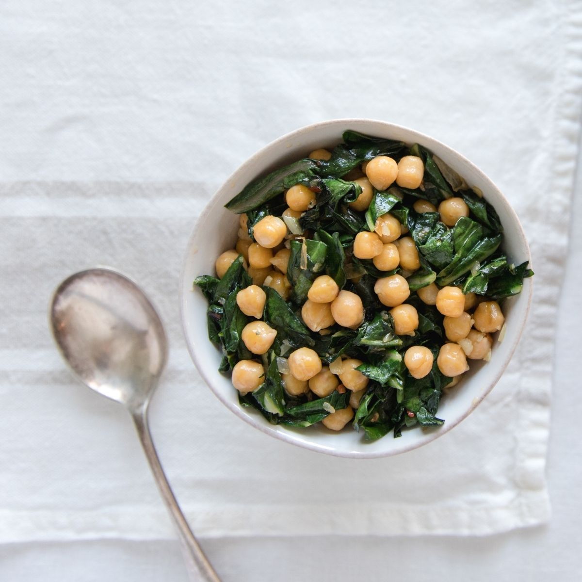 Top-down view of organic chickpeas with sautéed greens in a white bowl, on a white cloth with a serving spoon