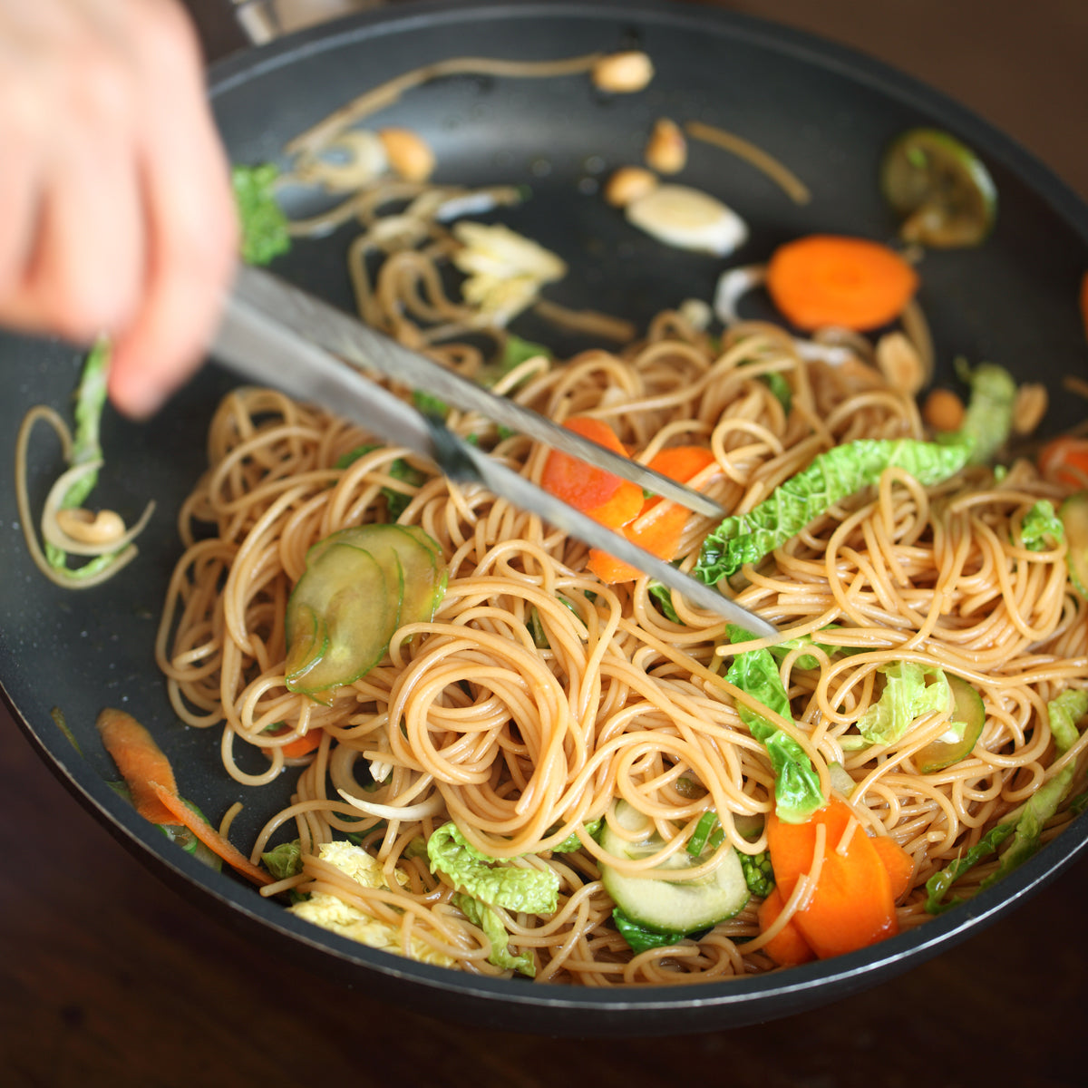 Person tossing organic gluten-free brown rice capellini with vegetables in a skillet